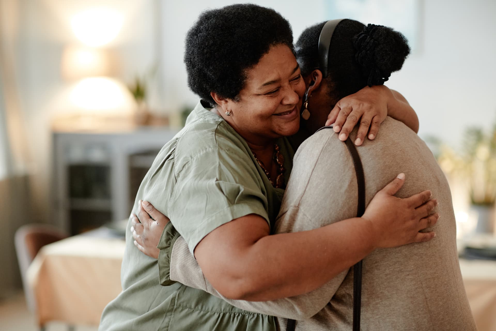 Two women embrace indoors, one smiling in a green shirt and the other wearing a beige cardigan.