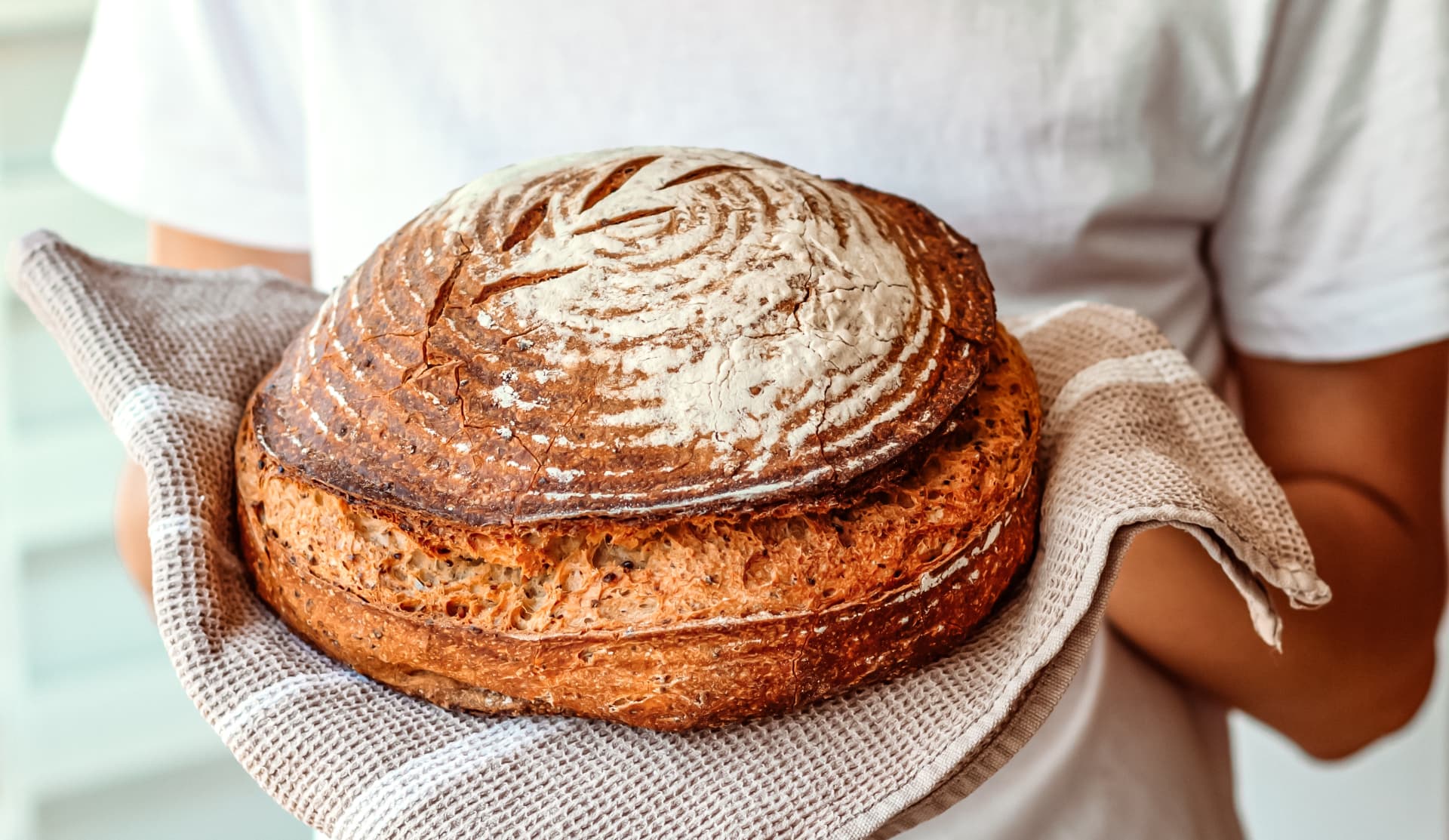 Hands holding a freshly baked round sourdough loaf with a flour-dusted, scored crust on a linen tea towel.