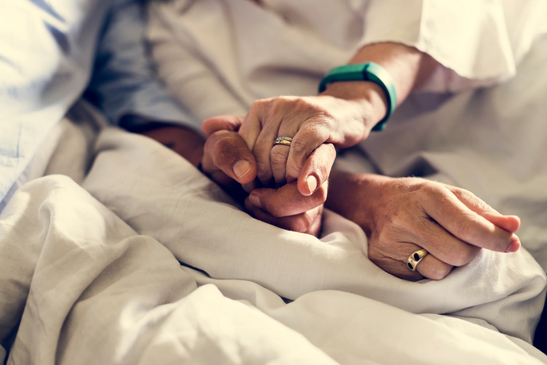Two older people's hands clasped together over cream-colored bed linens, one hand resting gently over the other, gold rings on both.