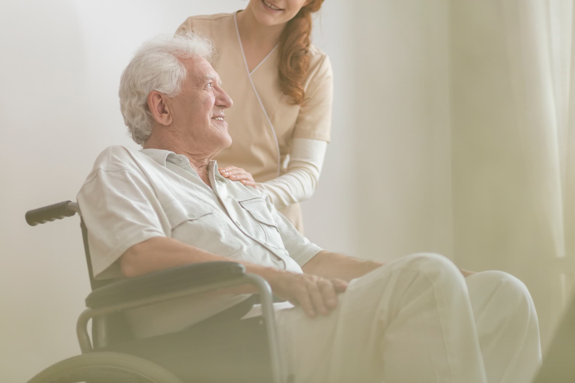 A smiling senior man in a wheelchair, a caregiver's hand resting gently on his shoulder