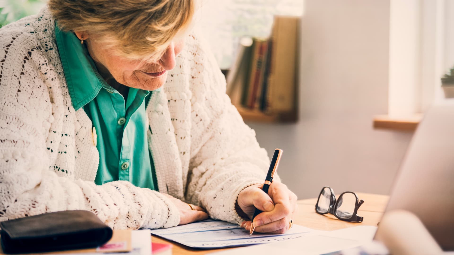 An older woman in a green shirt and cream cardigan writes on a form at a desk, with glasses beside her and a laptop nearby.
