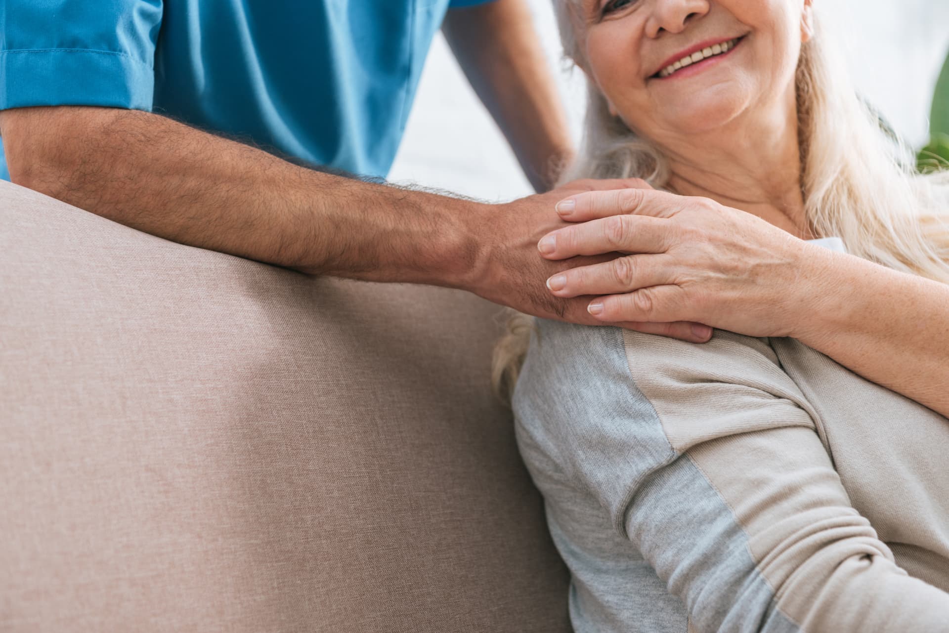 A smiling older woman with long white hair sits on a sofa while a man in blue medical scrubs beside her holds her hand in both of his.