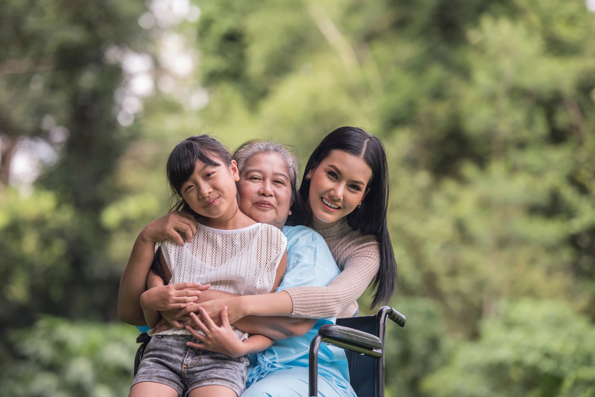 Three generations of a family embracing outdoors: a grandmother in a wheelchair hugged by her daughter and granddaughter, green trees behind them.