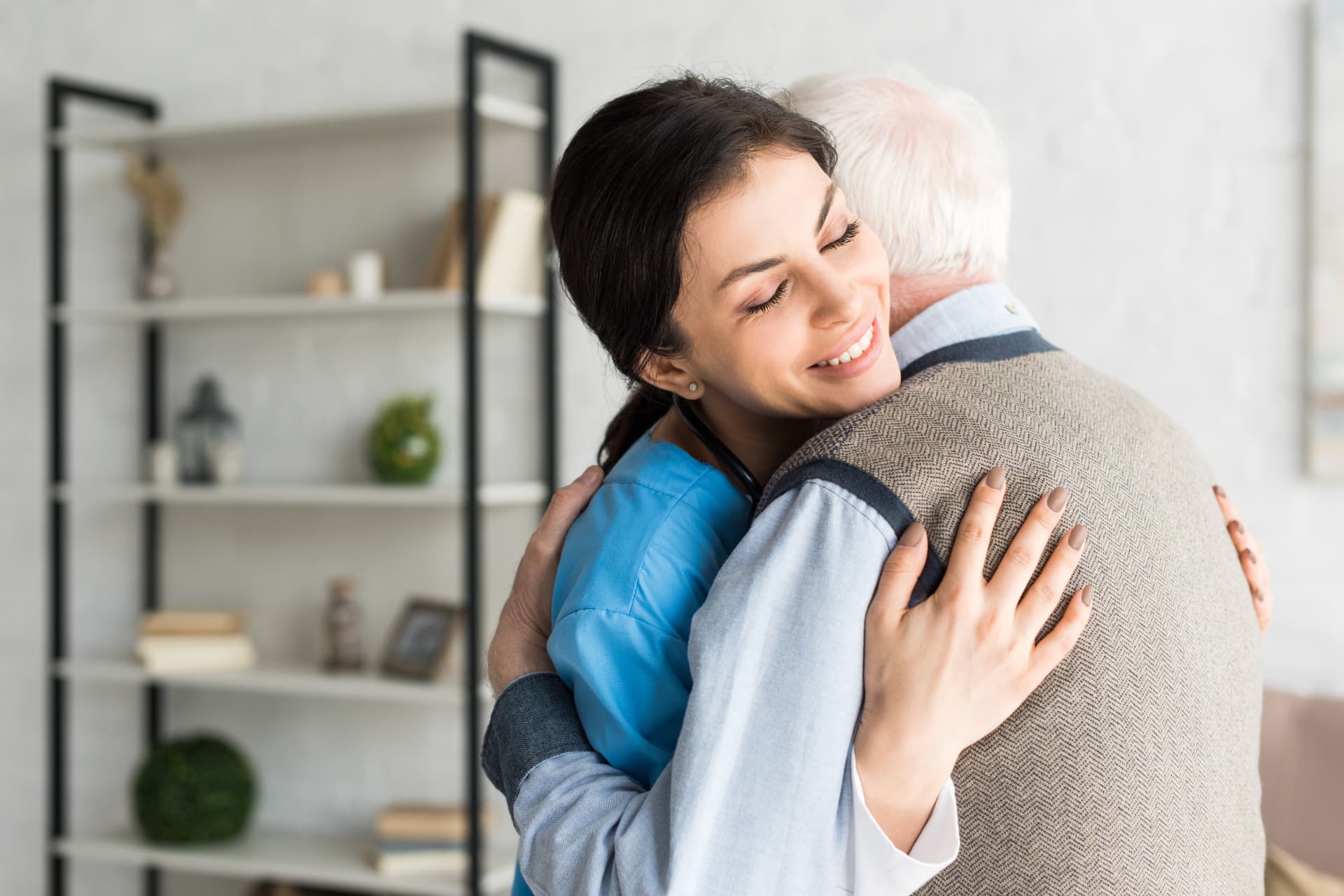 A young woman in blue medical scrubs closes her eyes as she hugs an older man in a tan sweater vest, his back to the camera, in a bright room with a bookshelf behind them.